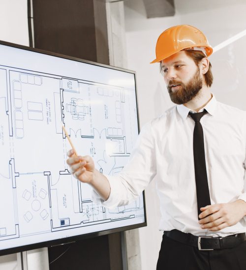 Young manager in the office with helmet Architect and engineer. Man shows plan of building. TV screen.